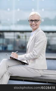 business, people and education concept - young smiling businesswoman in glasses with notepad over office building