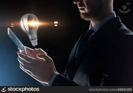 business, people and future technology concept - close up of businessman with transparent tablet pc computer and lightbulb projection over black background. close up of businessman with transparent tablet pc