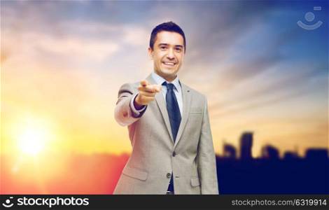 business, people and office concept - happy smiling businessman in suit pointing at you over city and sun light background. smiling businessman pointing at you over city