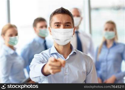 business, people and pandemic concept - businessman wearing face protective medical mask for protection from virus disease pointing finger to camera at office. businessman in mask pointing finger at office