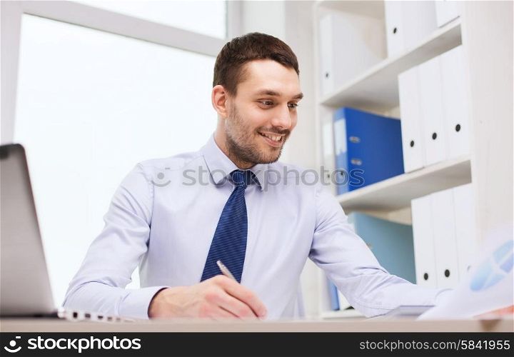 business, people and paper work concept - smiling businessman with laptop and papers writing in office