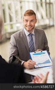 business, people and statistics concept - happy smiling young businessman with graph on clipboard meeting with colleague at office