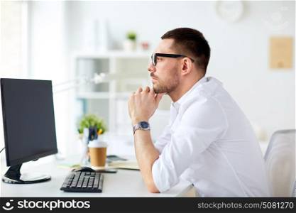 business, people and technology concept - businessman in glasses sitting at office computer and thinking. businessman in glasses sitting at office computer