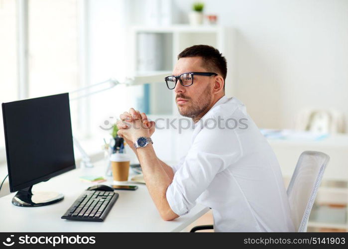 business, people and technology concept - businessman in glasses sitting at office computer and thinking. businessman in glasses sitting at office computer