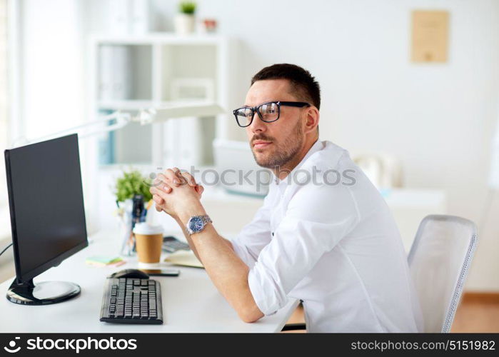 business, people and technology concept - businessman in glasses sitting at office computer and thinking. businessman in glasses sitting at office computer