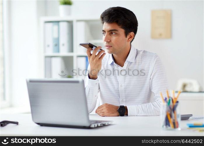 business, people and technology concept - businessman with laptop computer using voice command recorder on smartphone at office. businessman using voice recorder on smartphone