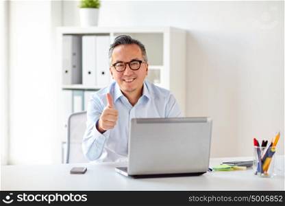 business, people and technology concept - happy smiling businessman in eyeglasses with laptop computer showing thumbs up at office. businessman with laptop showing thumbs up