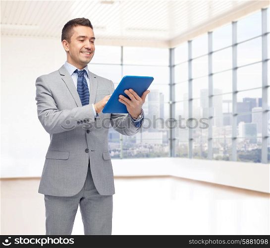 business, people and technology concept - happy smiling businessman in suit holding tablet pc computer over office room and window with city view background