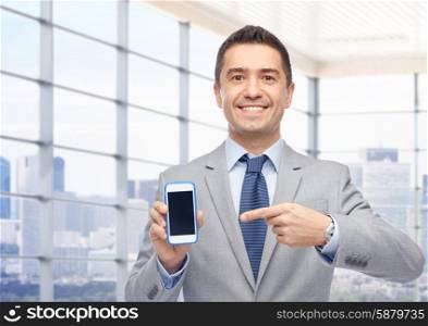 business, people and technology concept - happy smiling businessman in suit showing smartphone black blank screen over city office window background