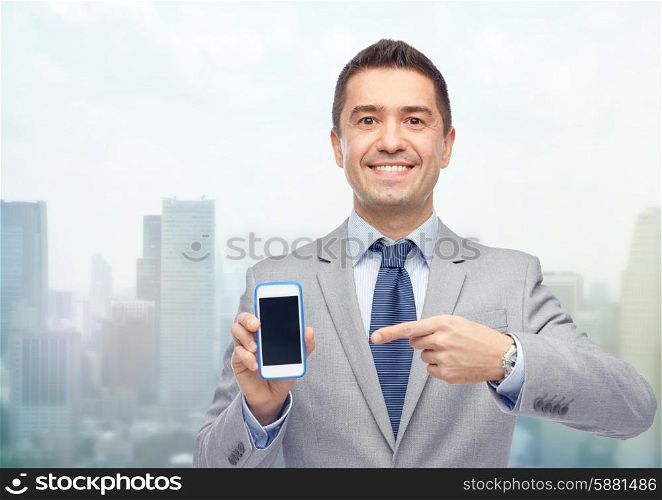 business, people and technology concept - happy smiling businessman in suit showing smartphone black blank screen over city background