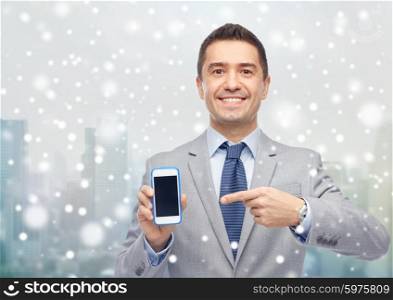 business, people and technology concept - happy smiling businessman in suit showing smartphone black blank screen over city background and snow effect