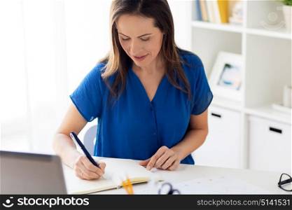 business, people and work concept - happy smiling woman with laptop computer working and writing to notebook at office or home. happy woman writing to notebook at office