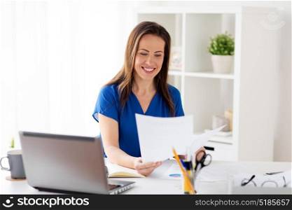 business, people and work concept - happy smiling woman with laptop computer working and writing to notebook at office or home. happy woman with papers and laptop at office