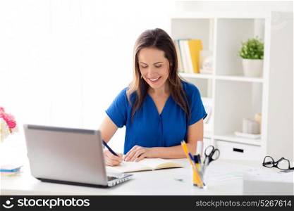 business, people and work concept - happy smiling woman with laptop computer working and writing to notebook at office or home. happy woman writing to notebook at office