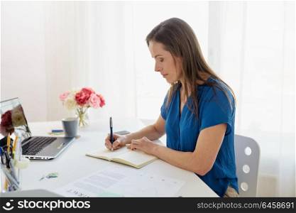 business, people and work concept - middle-aged woman with laptop computer working and writing to notebook at office or home. woman writing to notebook at office