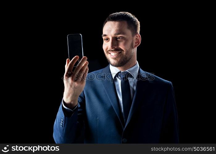 business, people, augmented reality and modern technology concept - smiling businessman in suit working with transparent smartphone over black background. businessman with transparent smartphone