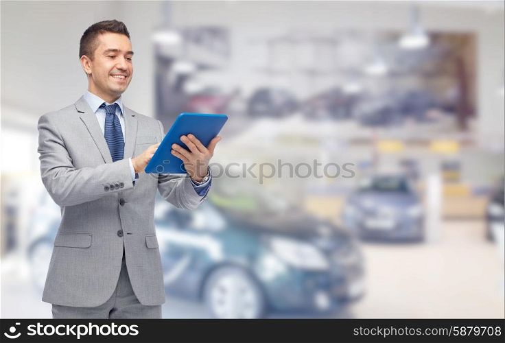 business, people, car sale and technology concept - happy smiling businessman in suit holding tablet pc computer over auto show or salon background