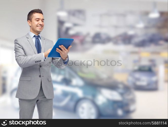 business, people, car sale and technology concept - happy smiling businessman in suit holding tablet pc computer over auto show or salon background