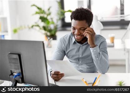 business, people, communication and technology concept - smiling african american businessman with papers and computer calling on smartphone at office. businessman calling on smartphone at office. businessman calling on smartphone at office