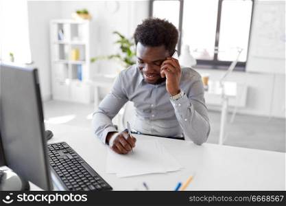 business, people, communication and technology concept - smiling african american businessman with papers and computer calling on smartphone at office. businessman calling on smartphone at office. businessman calling on smartphone at office