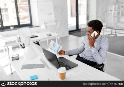 business, people, communication and technology concept - smiling african american businessman with papers and laptop computer calling on smartphone at office. businessman calling on smartphone at office. businessman calling on smartphone at office