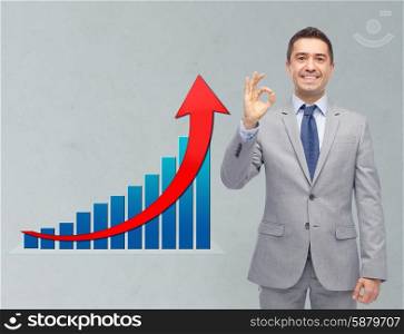 business, people, economics and financial success concept - happy smiling businessman in suit ok hand sign with growth chart over gray background