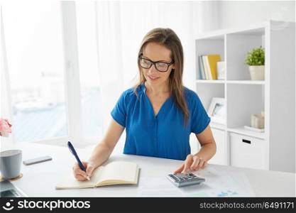 business, people, finance and accounting concept - happy woman with calculator counting and writing to notebook at office. happy woman with calculator and notebook at office. happy woman with calculator and notebook at office