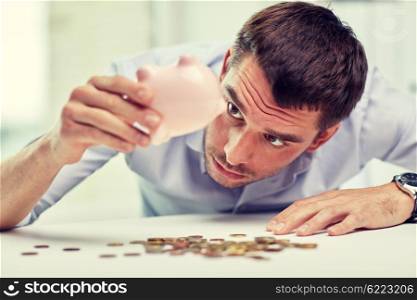 business, people, finances, crisis and money saving concept - businessman shaking piggy bank and getting coins at office