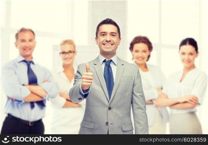 business, people, gesture and office concept - happy businessman with team over office room background showing thumbs up