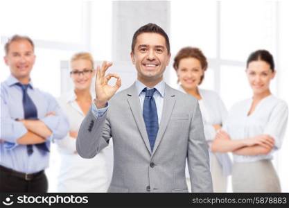 business, people, gesture and success concept - happy smiling businessman in suit with team over office room background showing ok hand sign