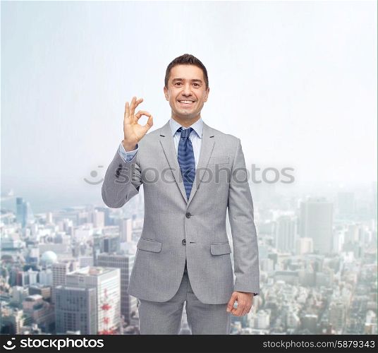 business, people, gesture and success concept - happy smiling businessman in suit ok hand sign over city background