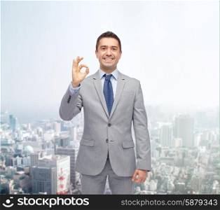business, people, gesture and success concept - happy smiling businessman in suit ok hand sign over city background