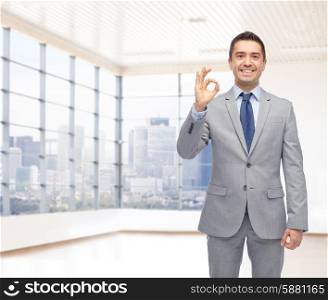 business, people, gesture and success concept - happy smiling businessman in suit ok hand sign over office room and window with city view background