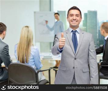business, people, gesture and success concept - happy smiling businessman with team over office room background showing thumbs up