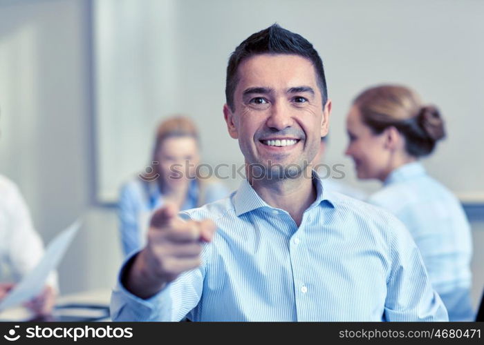 business, people, gesture and teamwork concept - smiling businessman pointing finger to you with group of businesspeople meeting in office