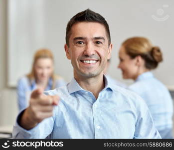 business, people, gesture and teamwork concept - smiling businessman pointing finger to you with group of businesspeople meeting in office