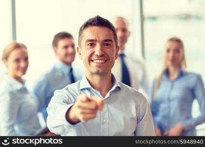 business, people, gesture and teamwork concept - smiling businessman pointing finger to you with group of businesspeople meeting in office