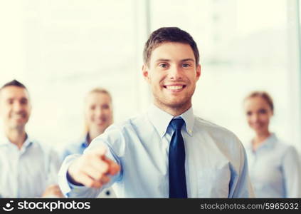 business, people, gesture and teamwork concept - smiling businessman pointing finger to you with group of businesspeople meeting in office