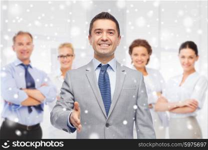 business, people, gesture, partnership and greeting concept - happy smiling businessman in suit with team giving hand for handshake over office room background and snow effect