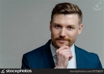 Business people. Headshot of handsome unshaved businessman or male entrepreneur in blue navy suit touching his chin and looking at camera thoughtfully while posing against grey studio background. Handsome thoughtful entrepreneur thinking about business and looking at camera while standing against grey background