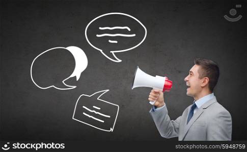 business, people, information and communication concept - happy businessman in suit speaking to megaphone and text bubbles over dark gray concrete wall background