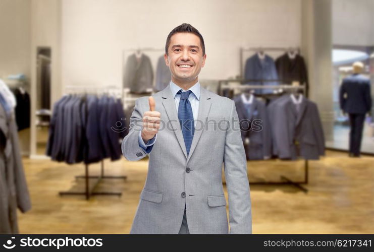 business, people, menswear, sale and clothes concept - happy smiling businessman in suit over clothing store background showing thumbs up