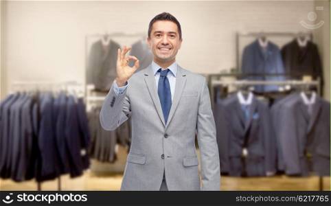 business, people, menswear, sale and clothes concept - happy smiling businessman in suit over clothing store background showing ok hand sign
