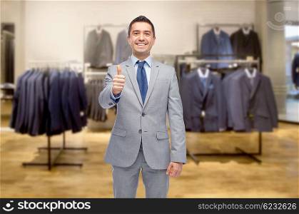 business, people, menswear, sale and clothes concept - happy smiling businessman in suit over clothing store background showing thumbs up