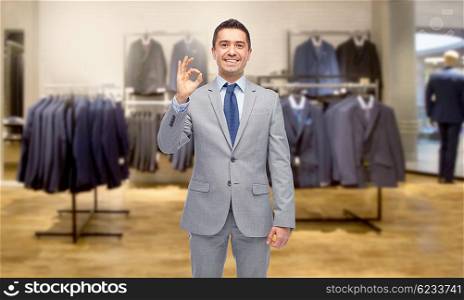 business, people, menswear, sale and clothes concept - happy smiling businessman in suit over clothing store background showing ok hand sign