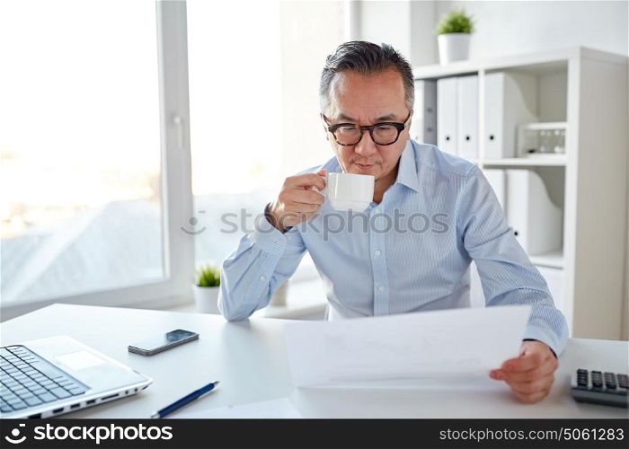 business, people, paperwork and technology concept - businessman reading paper and drinking coffee at office. businessman with papers drinking coffee at office