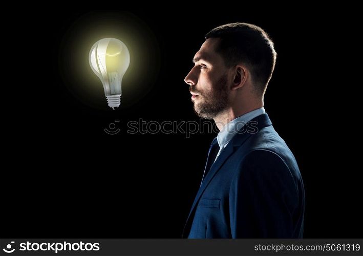 business, people, startup idea, inspiration and technology concept - businessman in suit looking at lightbulb over black background. businessman looking at lightbulb over black
