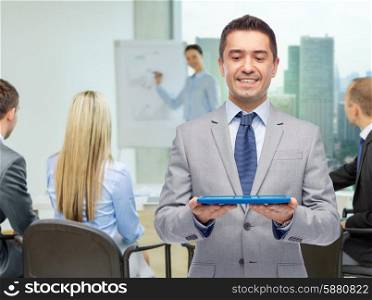 business, people, teamwork and technology concept - happy smiling businessman in suit holding tablet pc computer over group of people and office room background