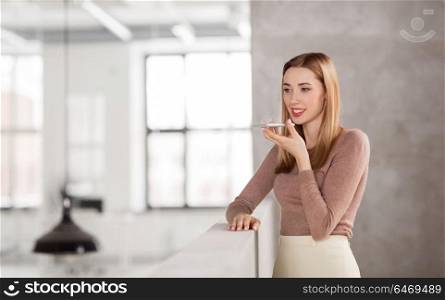business people, technology and corporate concept - happy smiling businesswoman using voice command recorder on smartphone at office. woman using voice recorder on smartphone at office
