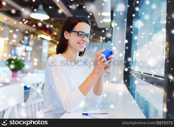 business, people, technology and lifestyle concept - smiling young woman in eyeglasses texting message with smartphone at cafe over snow effect
